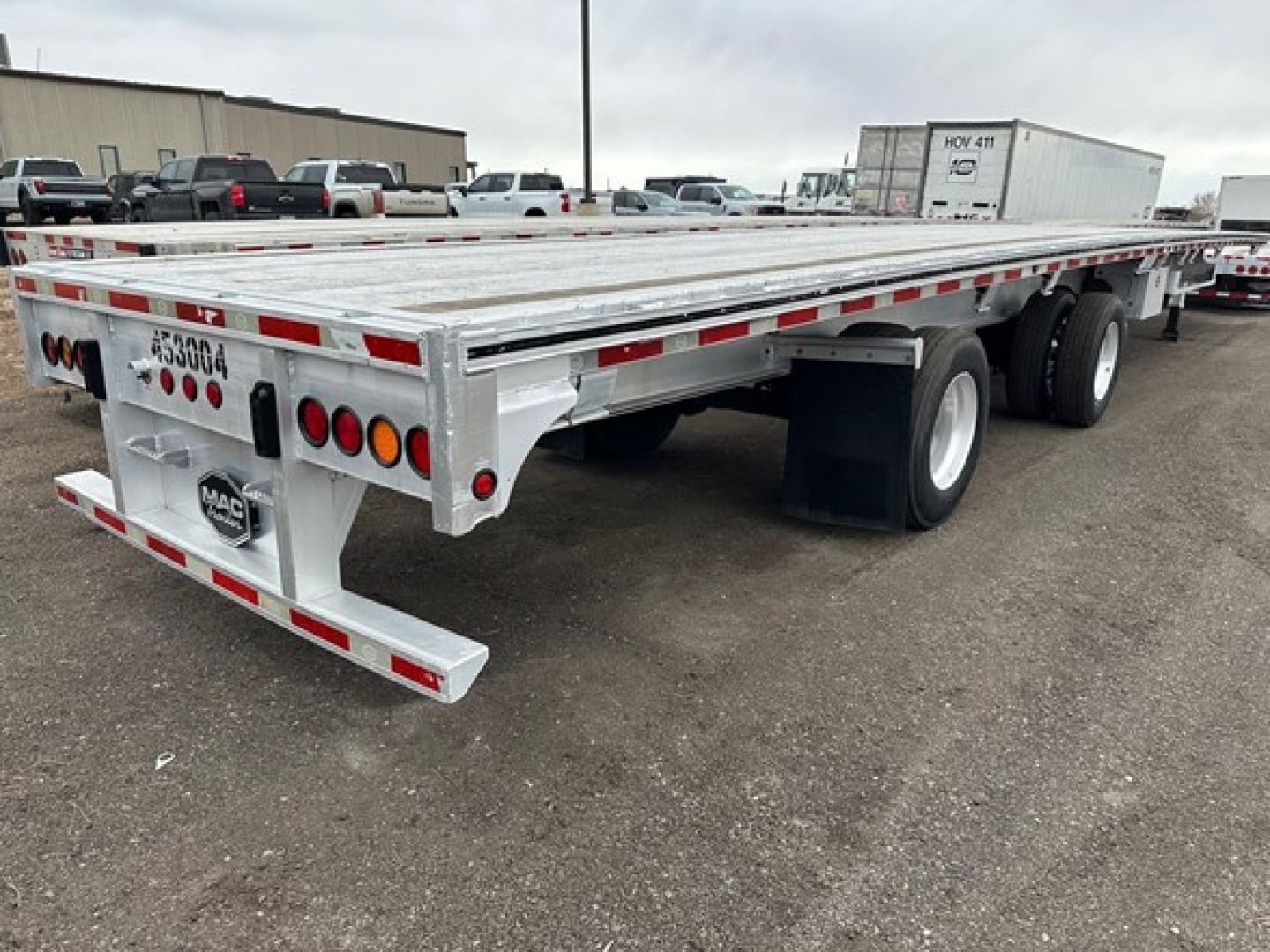 Flatbed trailer showing aluminum deck and dual rear axles with taillights. Visible text includes MAC trailer logo and rear certification plate; U-bolts and reflective tape along sides.