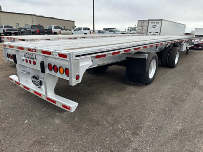 Flatbed trailer showing aluminum deck and dual rear axles with taillights. Visible text includes MAC trailer logo and rear certification plate; U-bolts and reflective tape along sides.