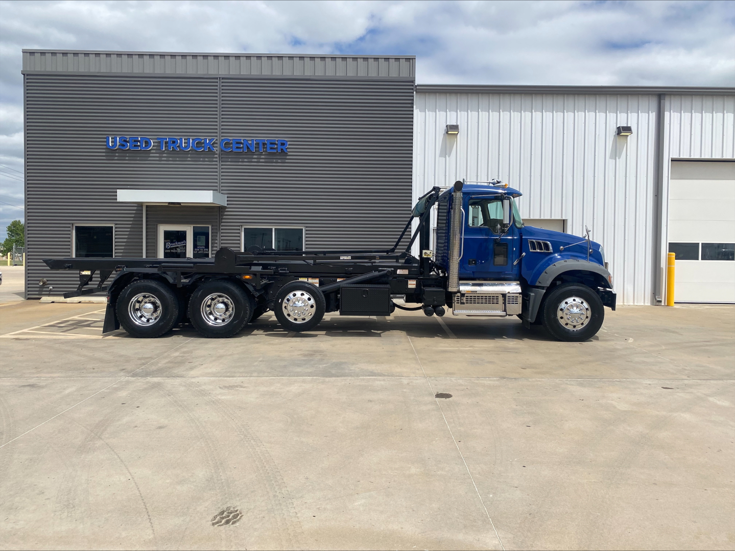 2019 Mack GR64F roll-off truck in blue cab with black chassis, side view showing multiple axles, parked in front of a Used Truck Center building with signage.