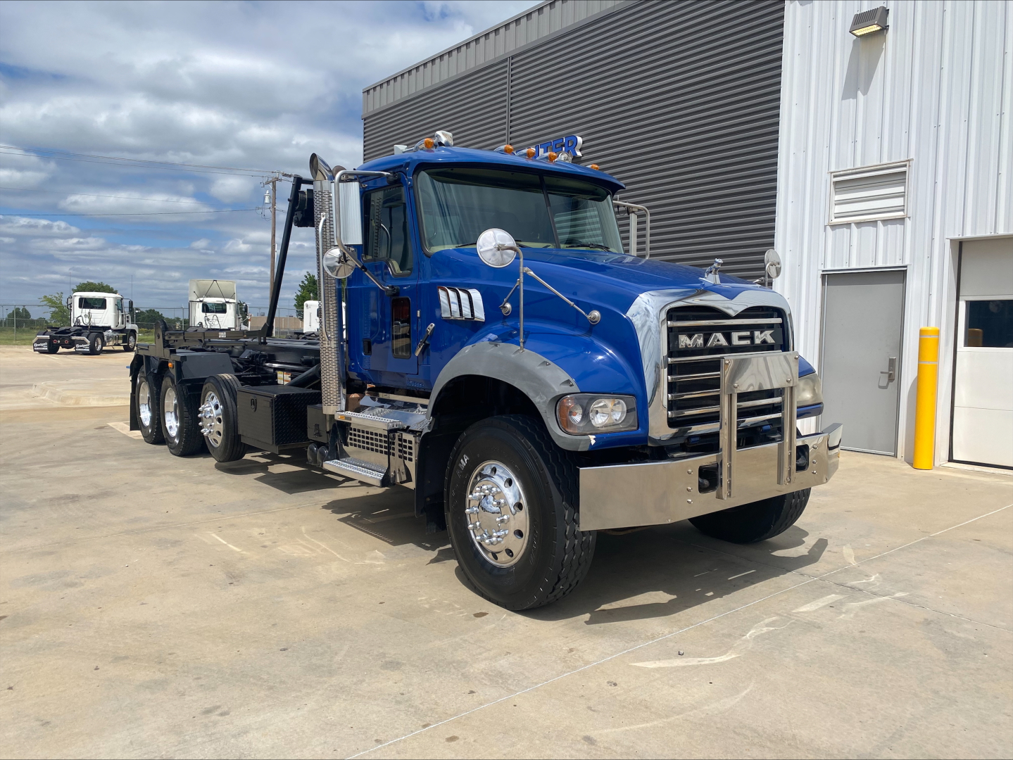 Blue Mack GR64F roll-off truck in front of a building showing chrome bumper, large grille with MACC logo, side mirrors, and dual rear axles; front fender and hood visible.