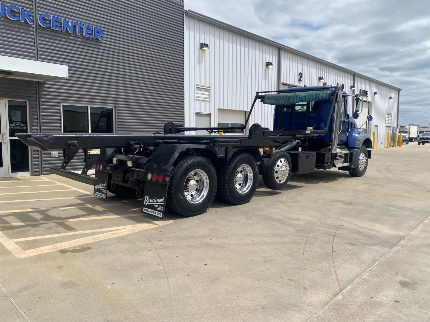 2019 Mack GR64F roll-off truck with blue cab and black flatbed, multiple axles, and rear wheels; visible text on rear mudflaps and building signage.