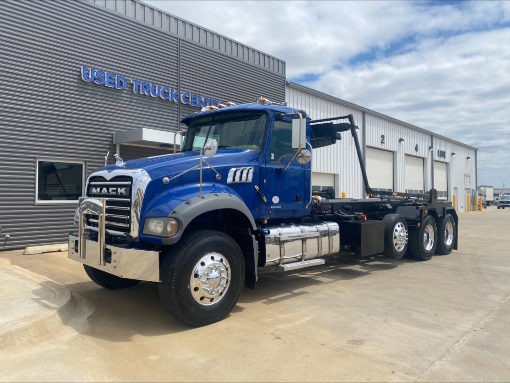 Mack GR64F truck in blue on lot showing front and side, log truck bed with extendable ramp, chrome bumper and wheel hubs.