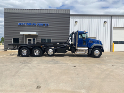 2019 Mack GR64F roll-off truck in blue cab with black chassis, side view showing multiple axles, parked in front of a Used Truck Center building with signage.