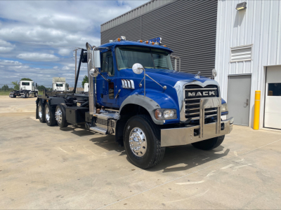 Blue Mack GR64F roll-off truck in front of a building showing chrome bumper, large grille with MACC logo, side mirrors, and dual rear axles; front fender and hood visible.