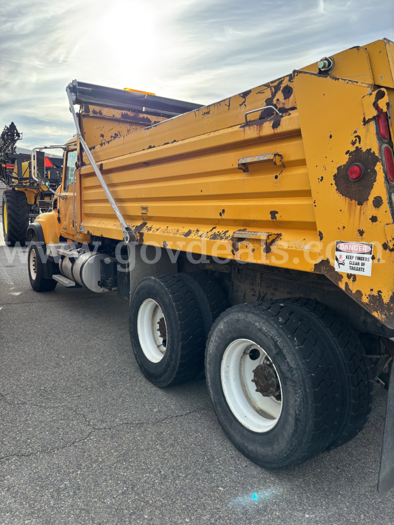 Dump truck rear view showing yellow Mack CV713 dump body with triple rear axles and: warning label on side, textured wear, hydraulic mechanism visible.
