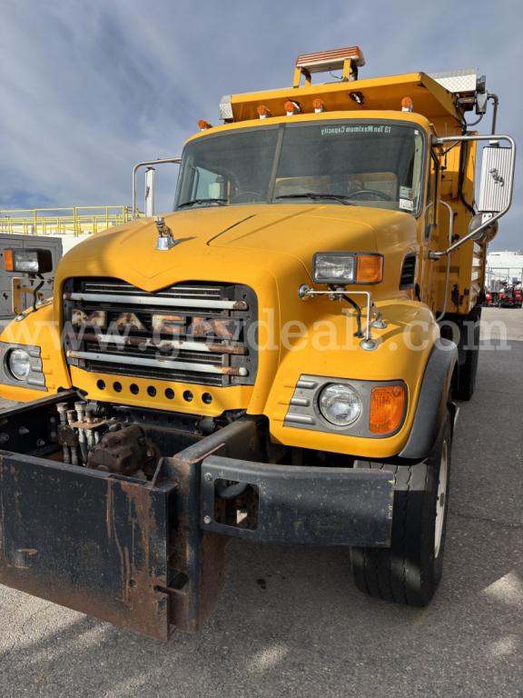 Yellow Mack CV713 dump truck front view with plow attachment and warning lights, grille, headlights, and cab mirrors visible.