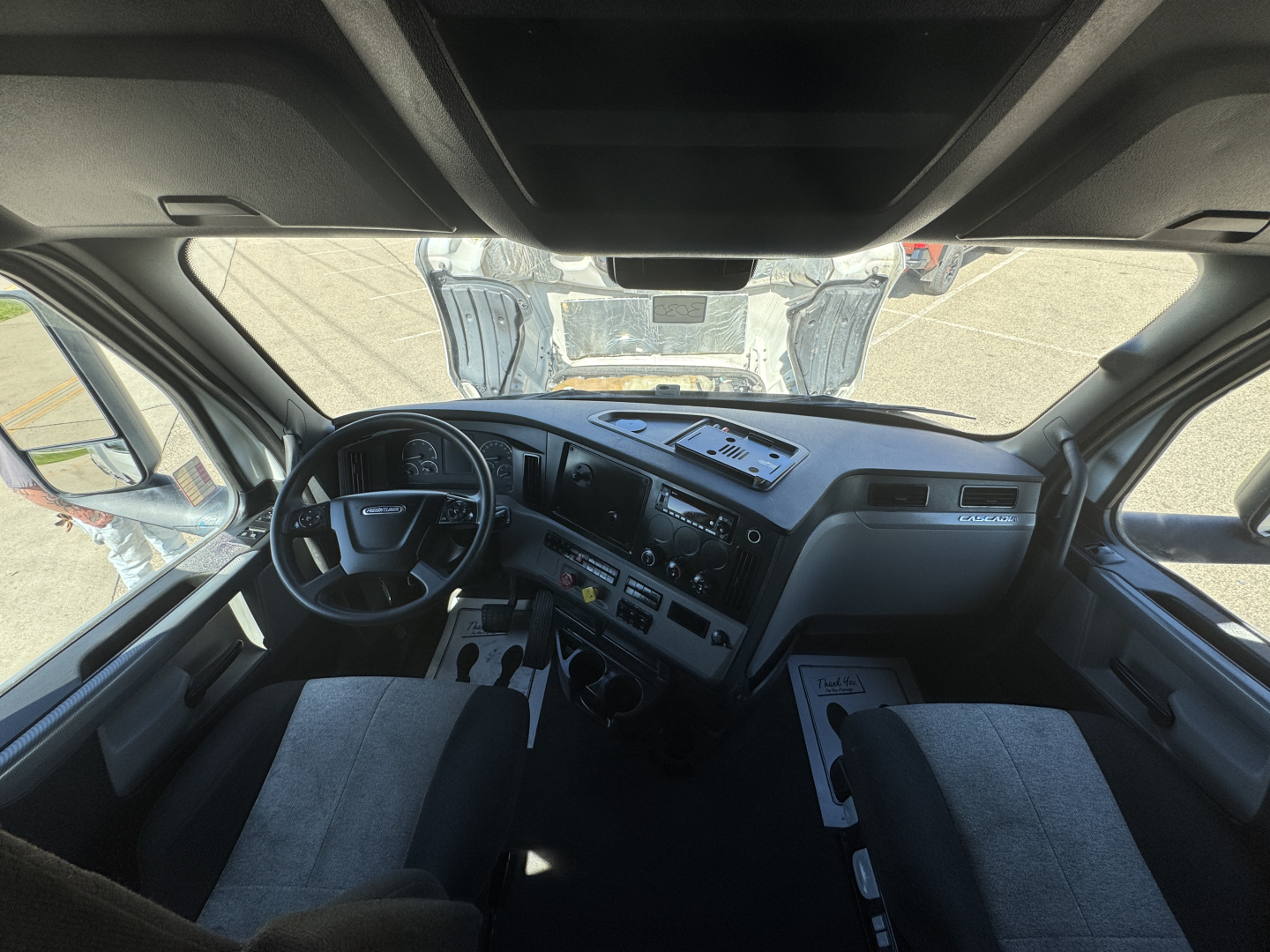 Cab interior of a Freightliner Cascadia truck with dashboard controls, steering wheel, and front seats visible through windshield.