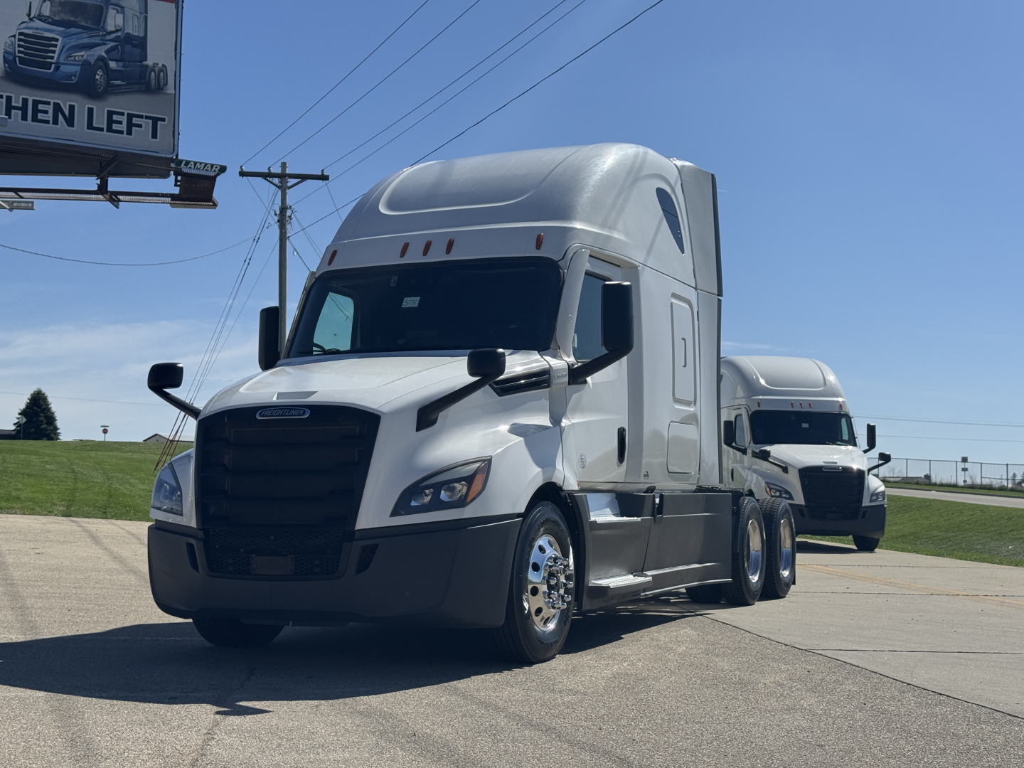 Freightliner Cascadia 126 semi-truck in a parking lot, white long-haul tractor with sleeper cab, front grille, dual headlights, side mirrors, and another truck in background.