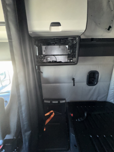 Truck cab interior showing dashboard cutaway and mounting hardware in the cabin of a Freightliner Cascadia sleeper; exposed metal panel with wiring and a small black in-cab console; vertical partition and floor area visible.