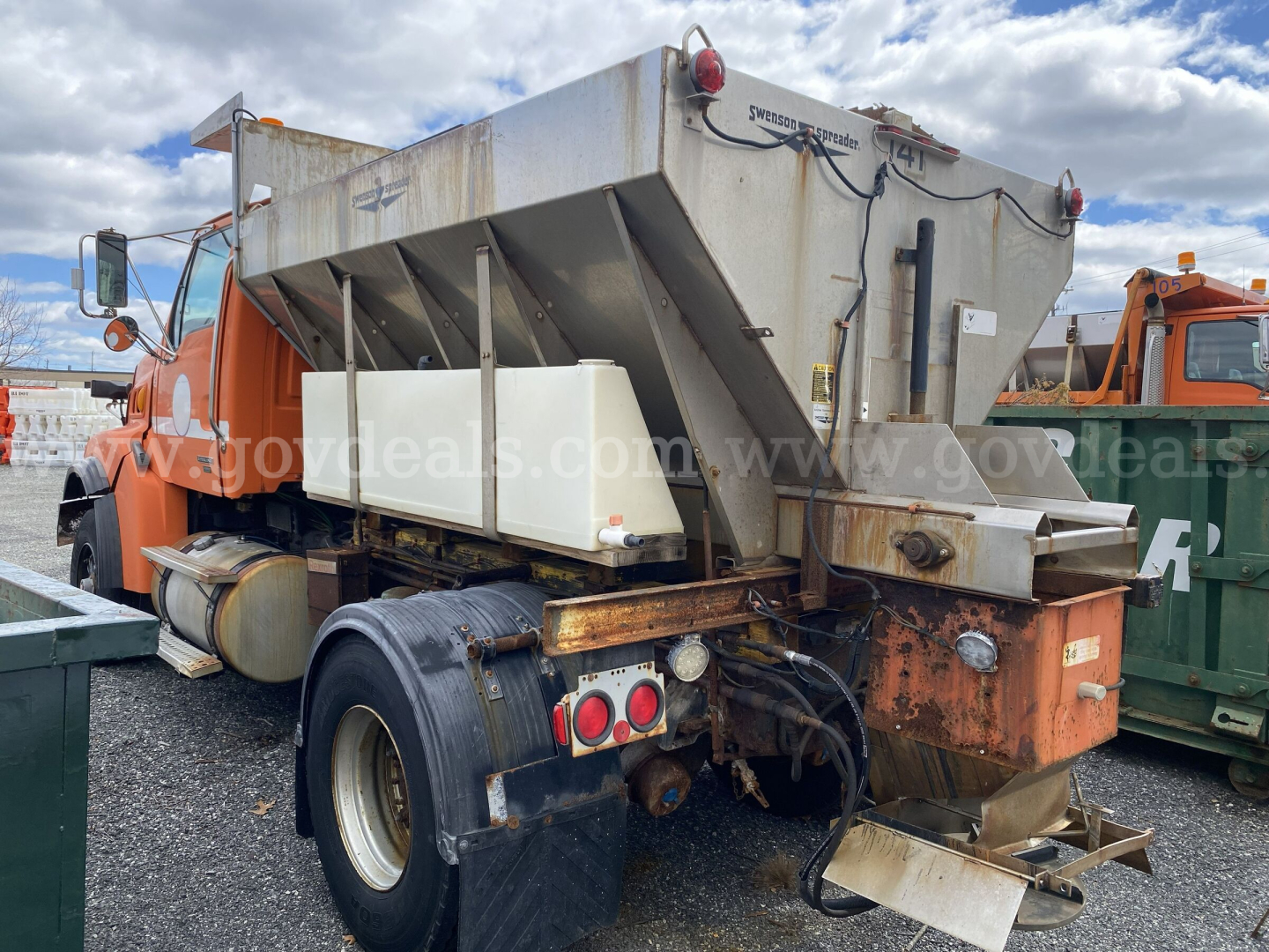 Dump truck rear view showing large aluminum hopper, tailgate, and dual rear wheels with worn second axle area; metal frame and taillight cluster visible.