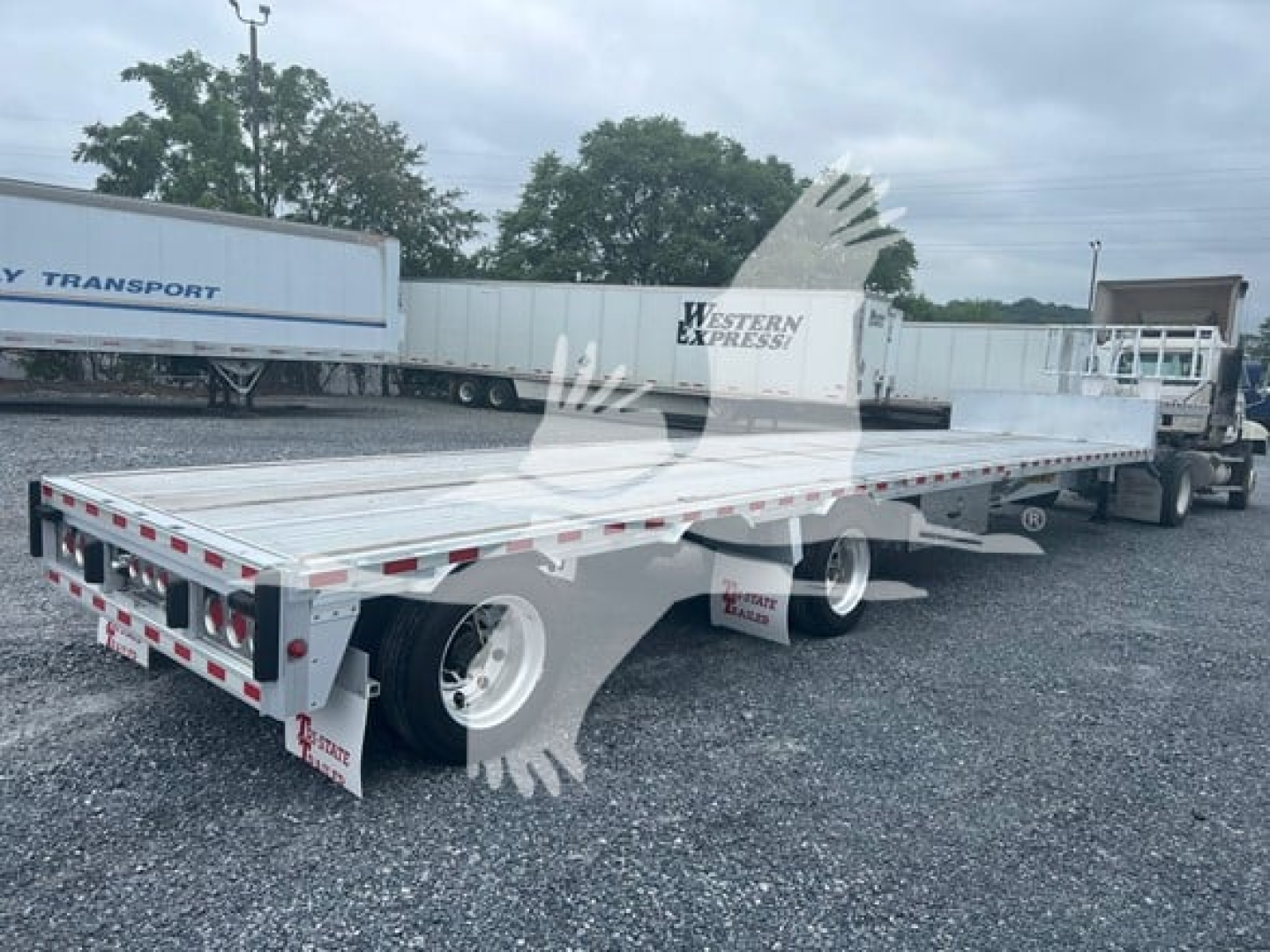 Flatbed trailer showing drop deck with metal surface and white wheels; rear taillights and reflective tape visible; label 'REITNOUER' on front side of trailer.