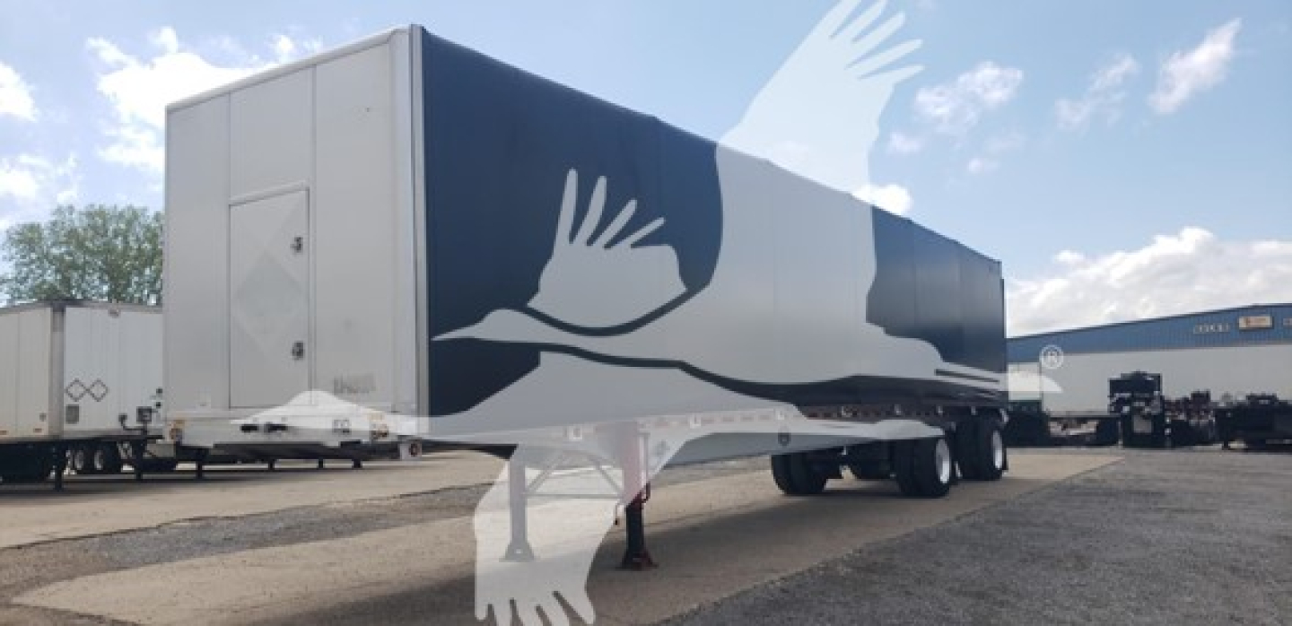 Curtainside trailer in a parking lot with blue and white graphics of birds in flight on the side. A white rear access door and front tractor disconnect area visible.