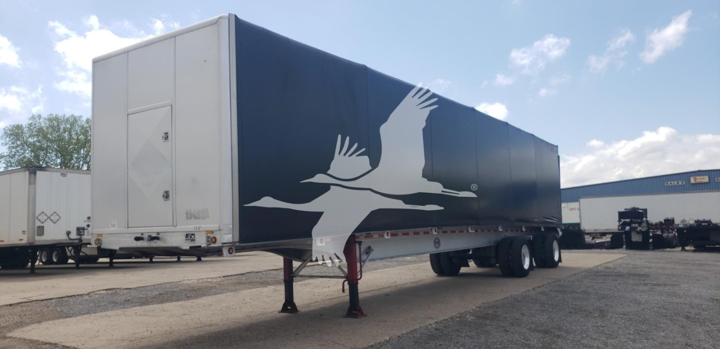 Flatbed trailer with curtain side showing a white bird graphic and blue exterior, front jack stands visible, serials not legible.