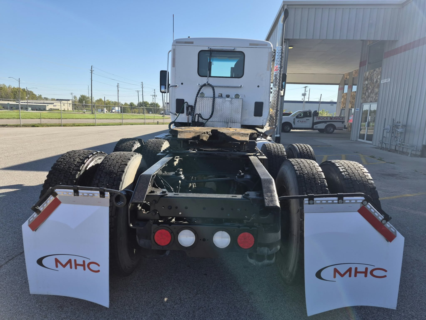 Rear view of a 2018 Kenworth T880 tractor chassis showing fifth wheel area, dual rear axles, dual fuel/air lines, MHC branded mud flaps on both sides.