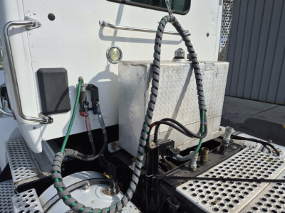 Kenworth T880 truck cab area showing fuel/DEF reservoir and engine bay components with hoses and metal diamond-plate step, side view.