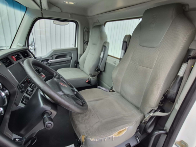 Kenworth T880 interior cab view showing driver's seat and dashboard from passenger side; fabric seat with wear on the edge; steering wheel, dashboard controls, and window provide view of modern truck interior.