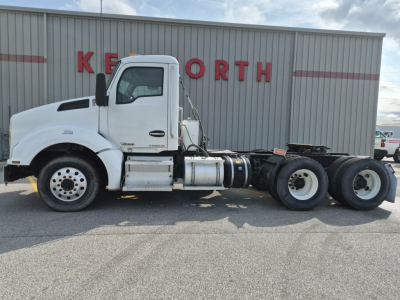 Truck side profile of a white 2018 Kenworth T880 conventional daycab chassis, showing cab, fuel tanks, and rear axles in front of a brick building.