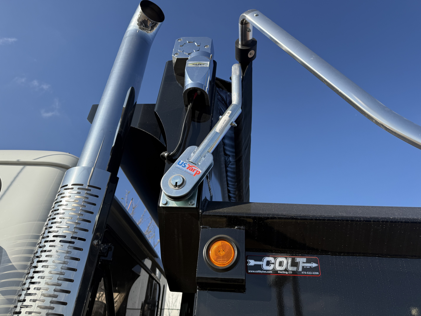 Mack PI64 dump truck tail area showing hydraulic hoist with chrome piston, black bed, orange indicator light, and COLT emblem. VIN plate not visible.