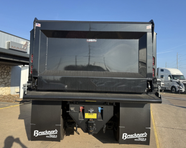 Mack PI64 dump truck dump bed rear view showing hydraulic system, tailgate, and brand stickers; back of truck with bumper and mud flaps.