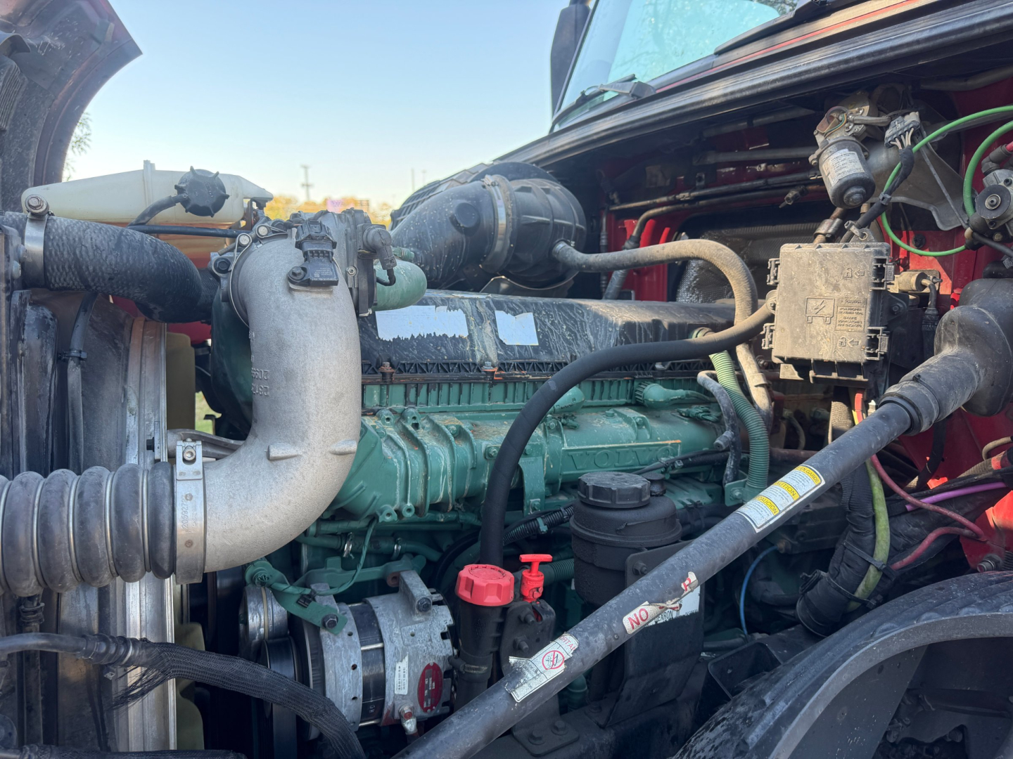 Volvo VNL64T760 engine bay showing diesel engine with green engine block, hoses, and battery; various labels and caps visible.