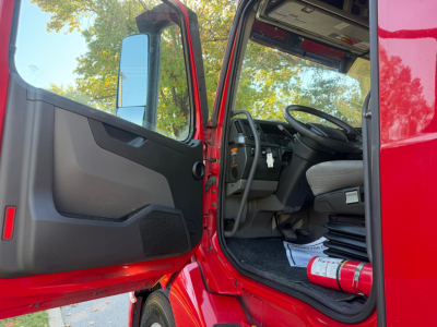 Cab interior of a red Volvo VNL truck with open door showing steering wheel and driver's seat, including a fire extinguisher on the floor and dashboard details.