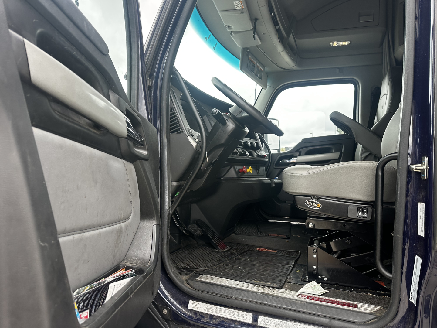 Kenworth T680 truck cab interior with steering wheel, dashboard, and gray driver seat; entry door open, dashboard console visible.