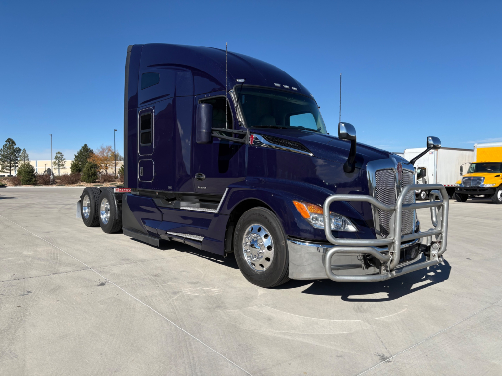 Conventional sleeper Kenworth T680 truck in purple/blue parked in lot, chrome bumper guard and grille, dual rear axles, side steps, showroom tires.
