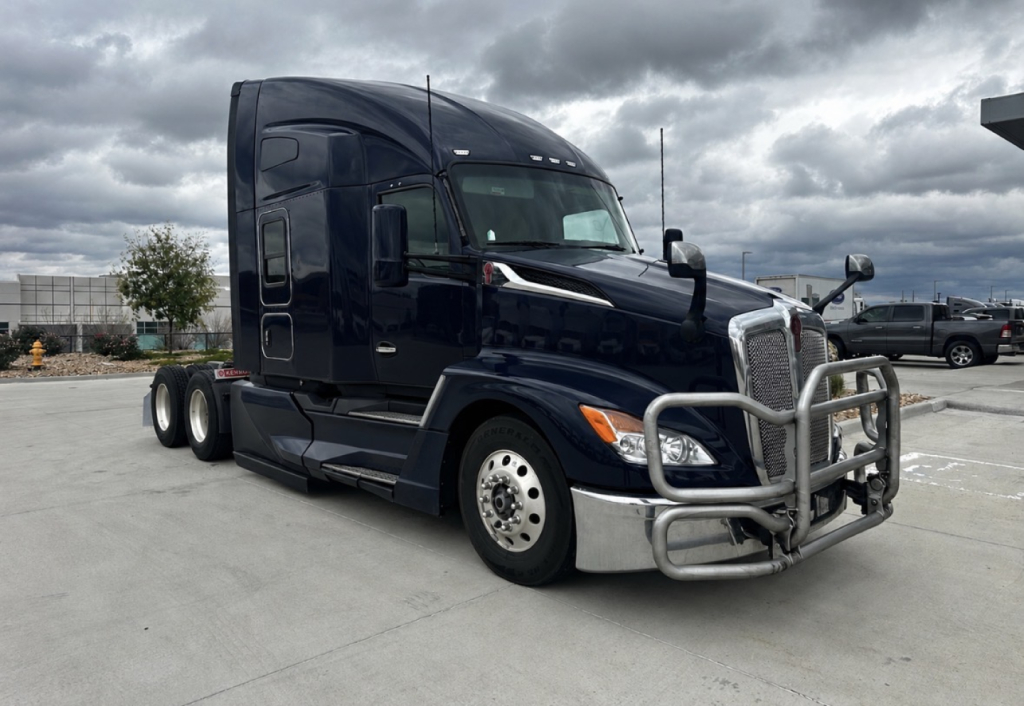 Kenworth T680 conventional sleeper truck in dark blue, front grille guard, chrome bumper, side mirrors, wheels, and dual rear axles visible.