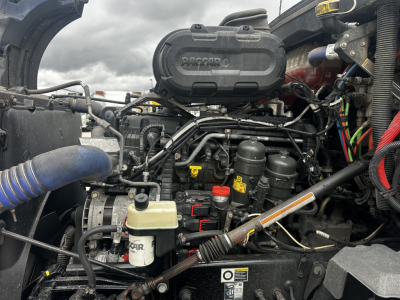 Kenworth T680 engine bay showing air intake, hoses, and various fluid reservoirs; visible components include an air filter housing labeled PACCAR, coolant/flush reservoirs, and wiring harnesses.