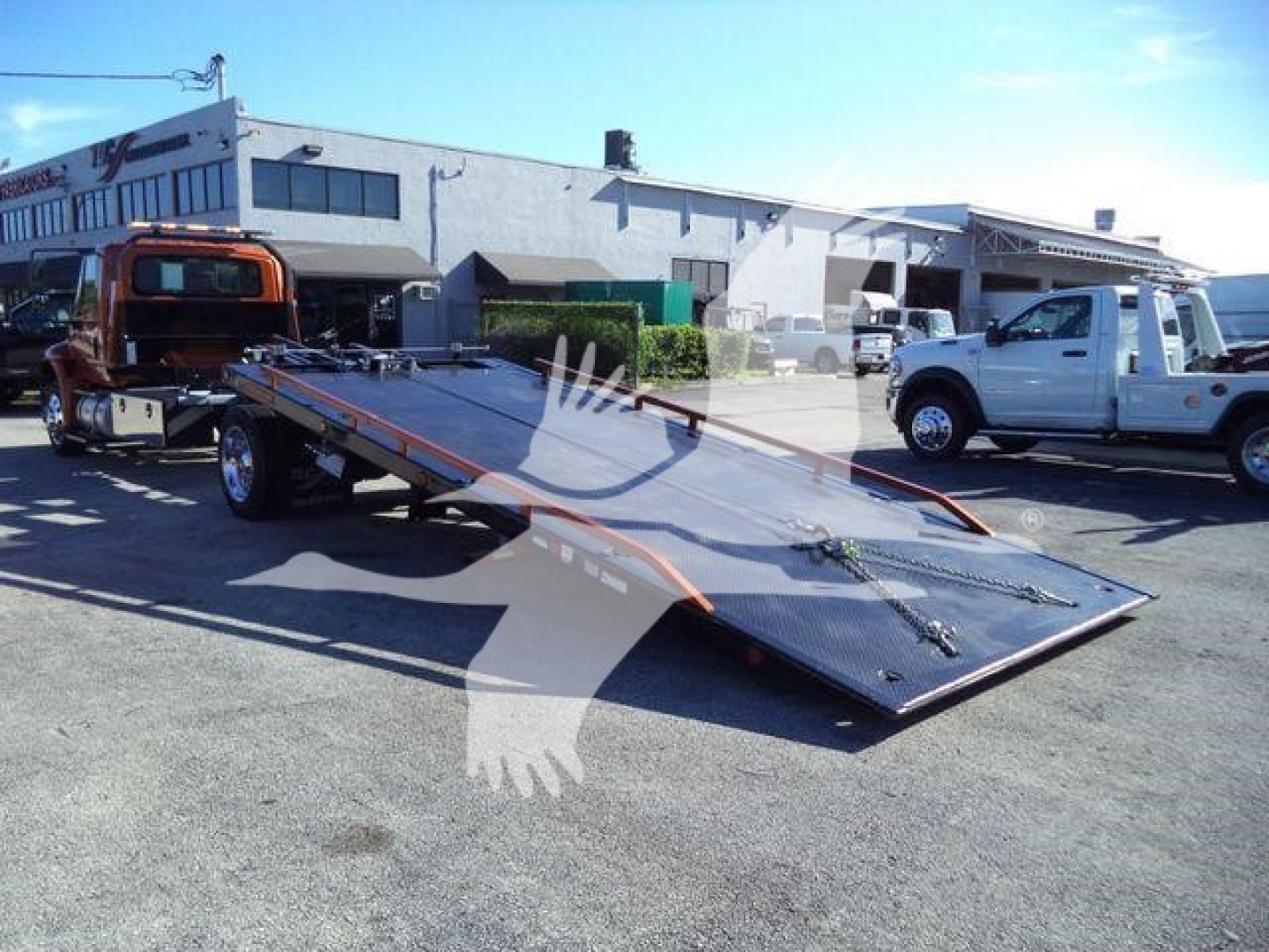 Flatbed tow truck loading plate with a white pickup truck and industrial warehouse in the background.