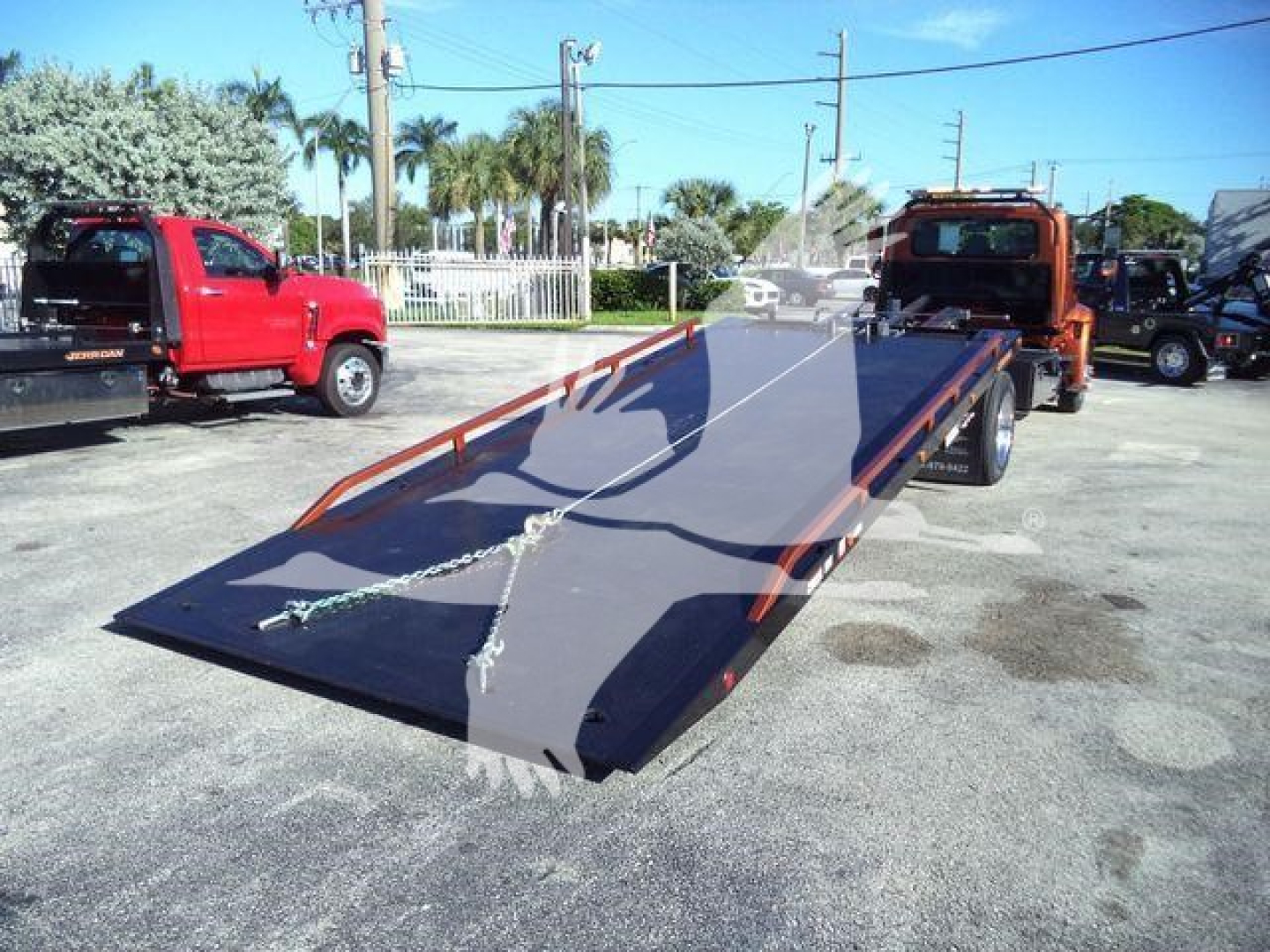 Flatbed tow truck in a dealership lot showing a long blue steel ramp flatbed with chains and a reflection of palm trees in the background.