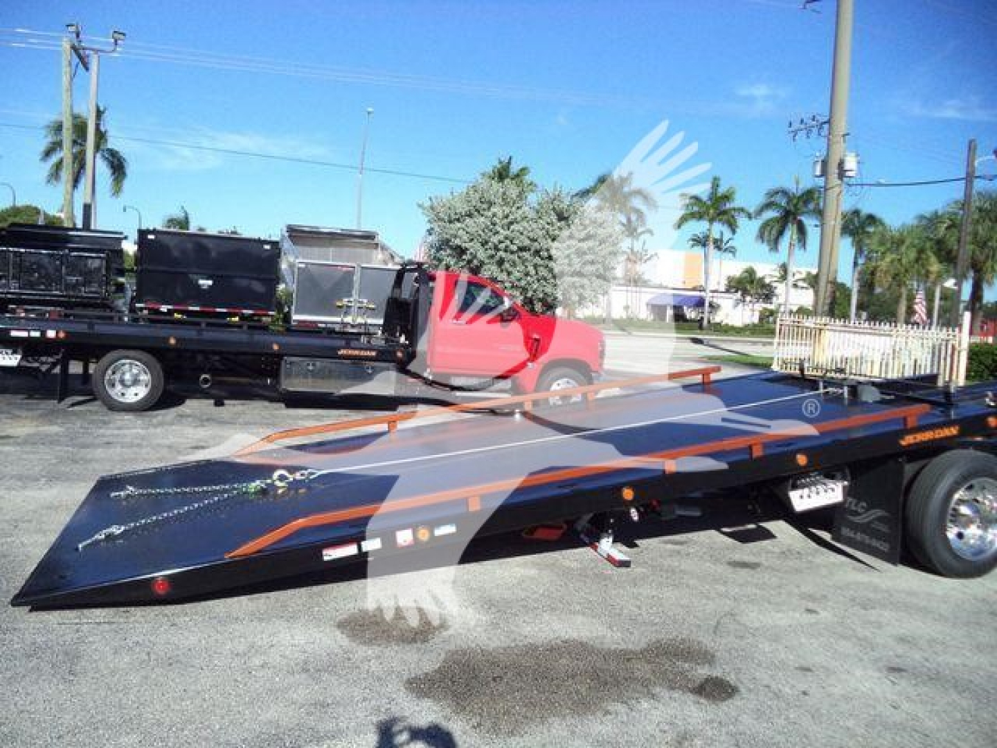 Truck bed in a raised position attached to a red International 4300 tow truck with a flatbed trailer, visible in a lot with palm trees.