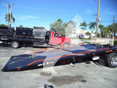 Truck bed in a raised position attached to a red International 4300 tow truck with a flatbed trailer, visible in a lot with palm trees.
