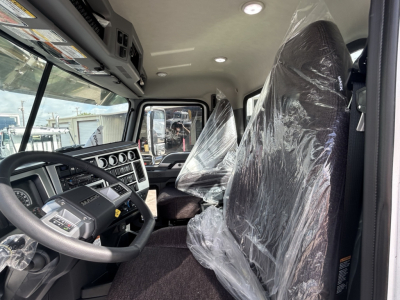Front cabin of a new Mack GR84FT truck showing steering wheel and driver's seat covered in plastic. Dashboard display with gauges and controls visible.