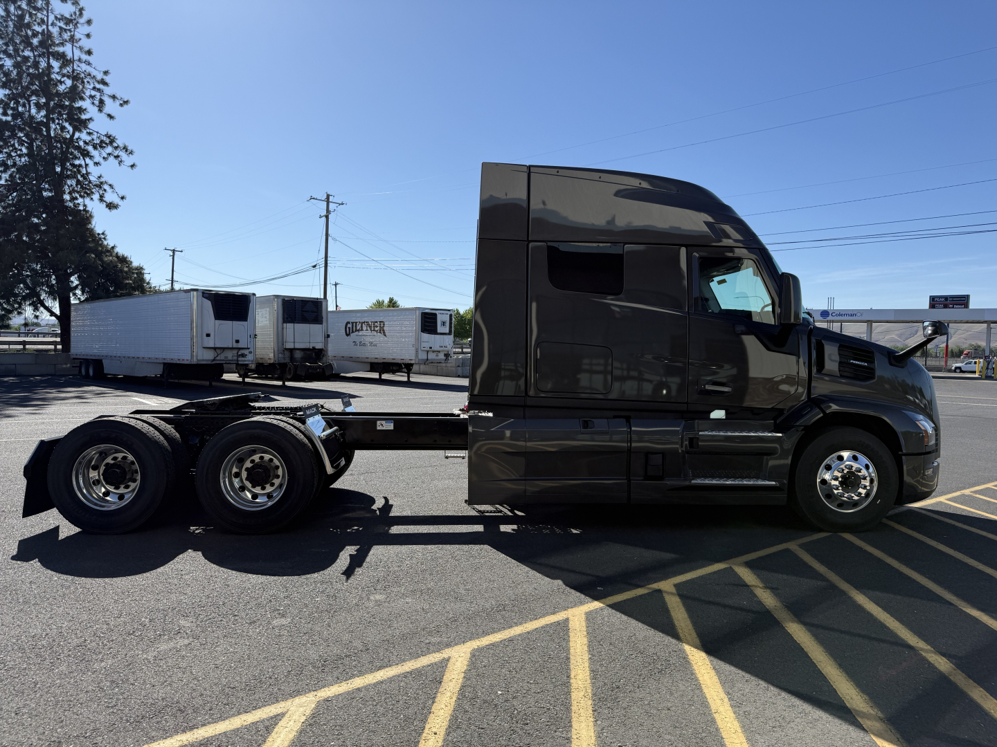 Side view of a black 2026 Mack PR64T64M conventional sleeper truck tractor with a long chassis and three rear axles showing. Visible text includes no explicit labels on the cab in the image.