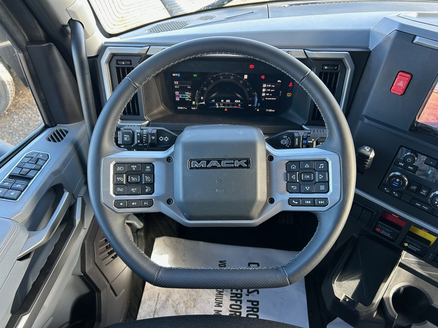 Mack PR64T64M truck interior showing steering wheel with Mack logo and digital instrument cluster, with controls on both sides of wheel.