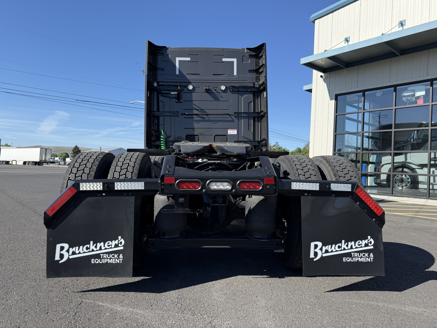 Rear view of a new Mack PR64T64M conventional sleeper truck with attached mudflaps reading Brackner's Truck & Equipment, dual rear wheels, and a fifth wheel hitch.