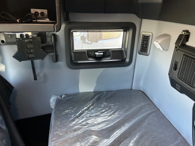 Cab interior of a 2026 Mack PR64T64M truck sleeper showing small window, overhead console, and seating area with protective plastic covering on the bed.
