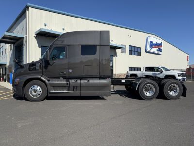 New Mack PR64T64M conventional sleeper tractor in dark gray, side view, with sleeper cab, dual rear axles, parked in lot outside dealership.