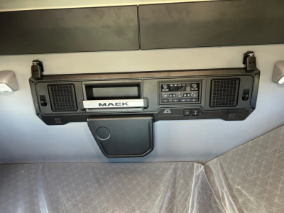 Dashboard control panel of a Mack truck. Center panel includes a 'MACK' badge, an infotainment style unit, two air vent grills on either side, and a storage compartment below. Looks like a modern cab interior.