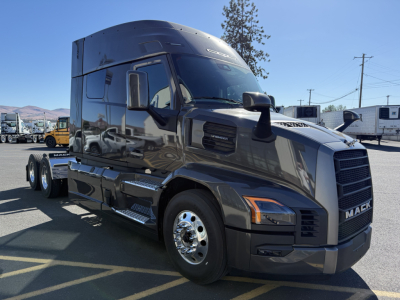 Mack PR64T64M tractor-trailer in dark gray showing cab with chrome wheels and side steps. Front grille badge reads MACK; diesel truck in lot.