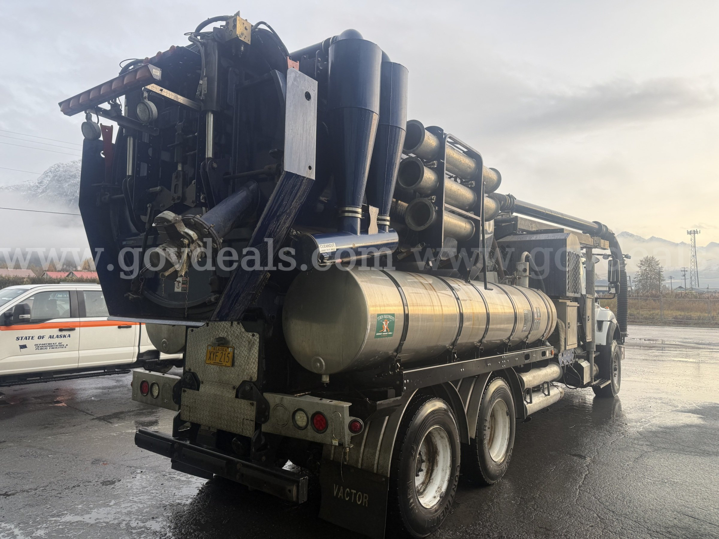 Rear view of a vacuum truck with large cylindrical tank and mounted hoses; shows various pipes, valves, and a towing hitch; faint watermark across image; no legible stickers readable.