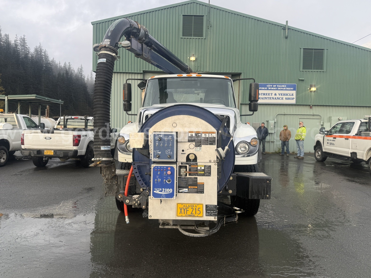 Front view of a 2013 International WorkStar 7500 with a large vacuum/loader unit mounted, showing hoses, control panel, and rear chute. Vehicle in yard with building in background.