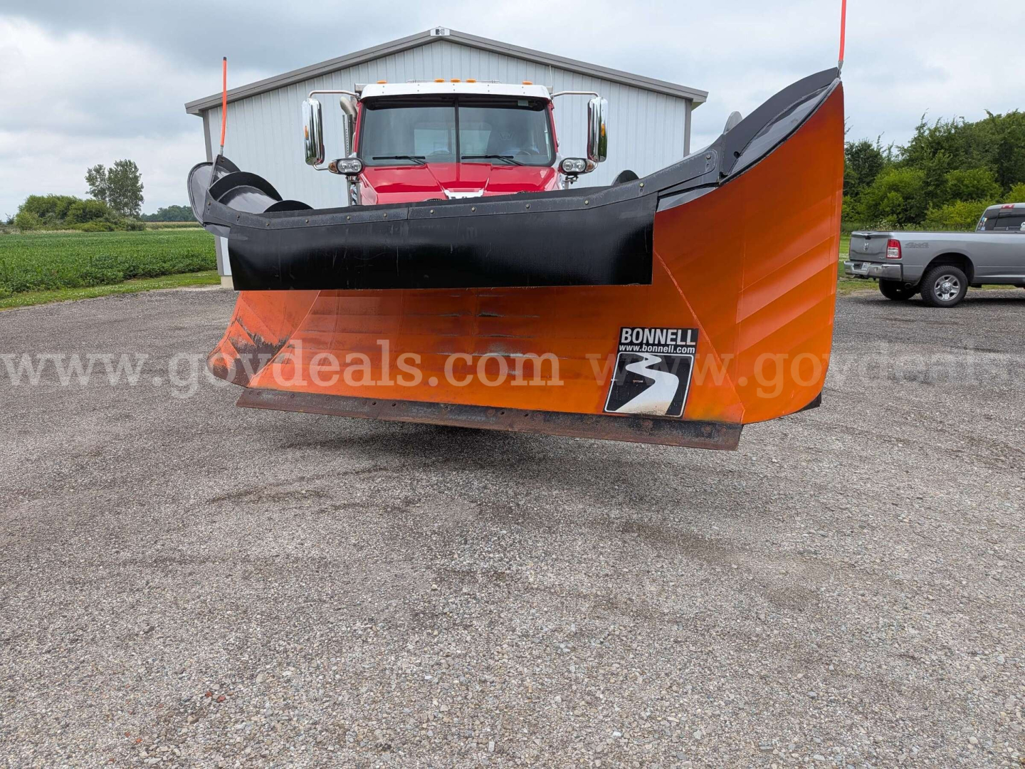 Snow plow blade attachment in front of a red truck, large orange and black Bonneville brand plow with white text and watermarked gears in background.