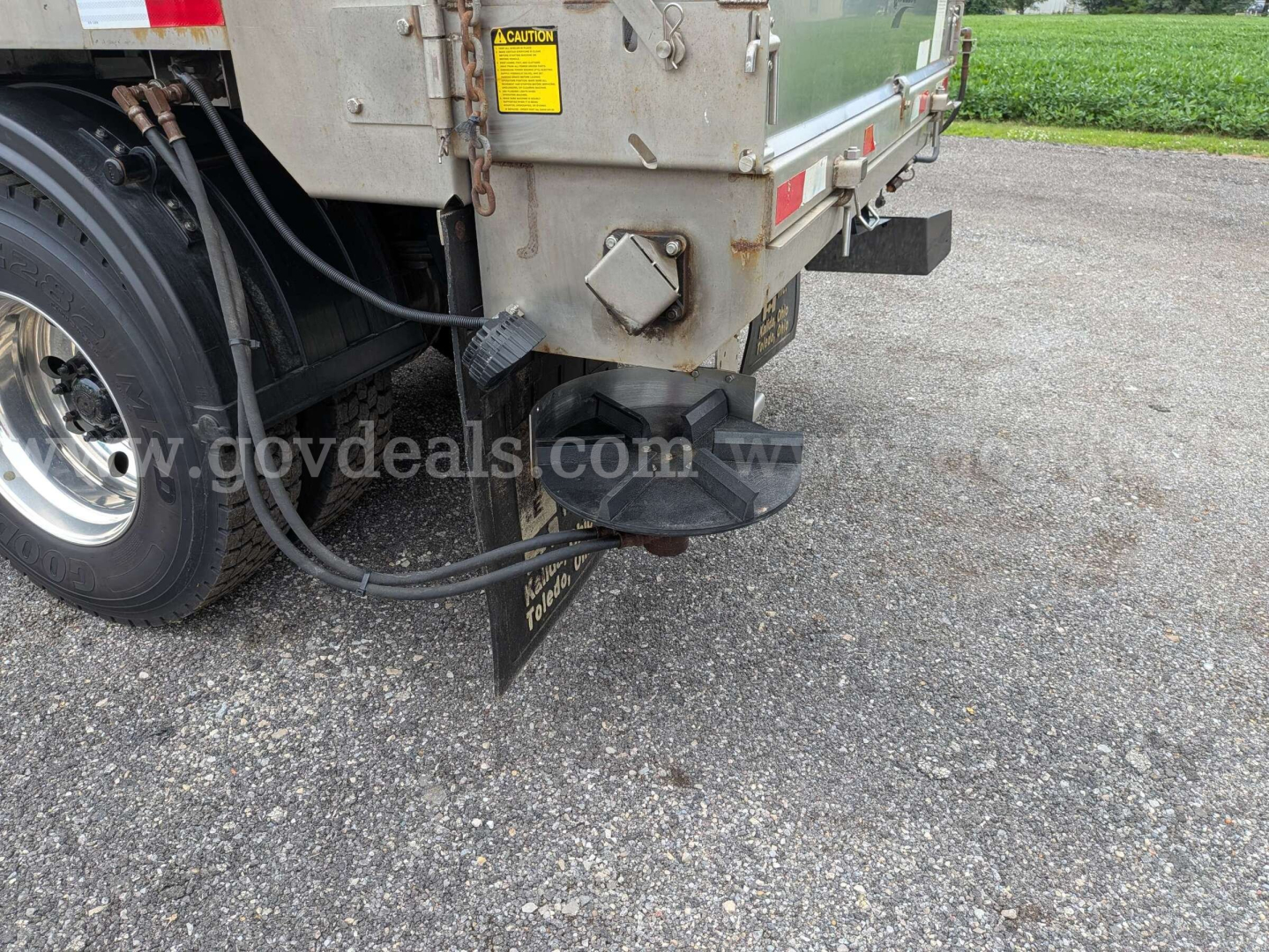 Rear left side of a 2015 Western Star 4700 dump/plow truck showing wheel, hydraulic hoses, and mounting bracket with caution sticker and a round black spare/ tool holder under the rear. Visible text includes caution label and straps.