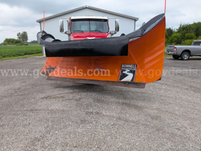 Snow plow blade attachment in front of a red truck, large orange and black Bonneville brand plow with white text and watermarked gears in background.