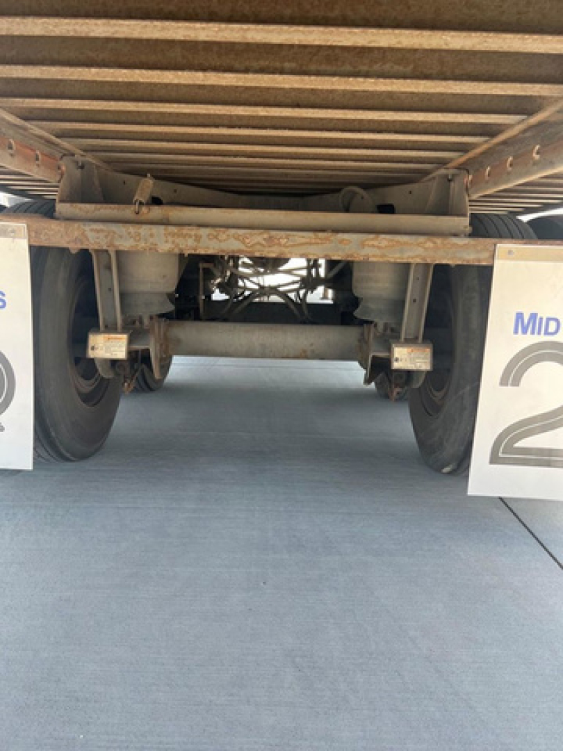 Underneath a utility van trailer showing rear axle assembly, leaf springs, and mud flaps with partial 'Mid' signage. Rust and metal frame visible.