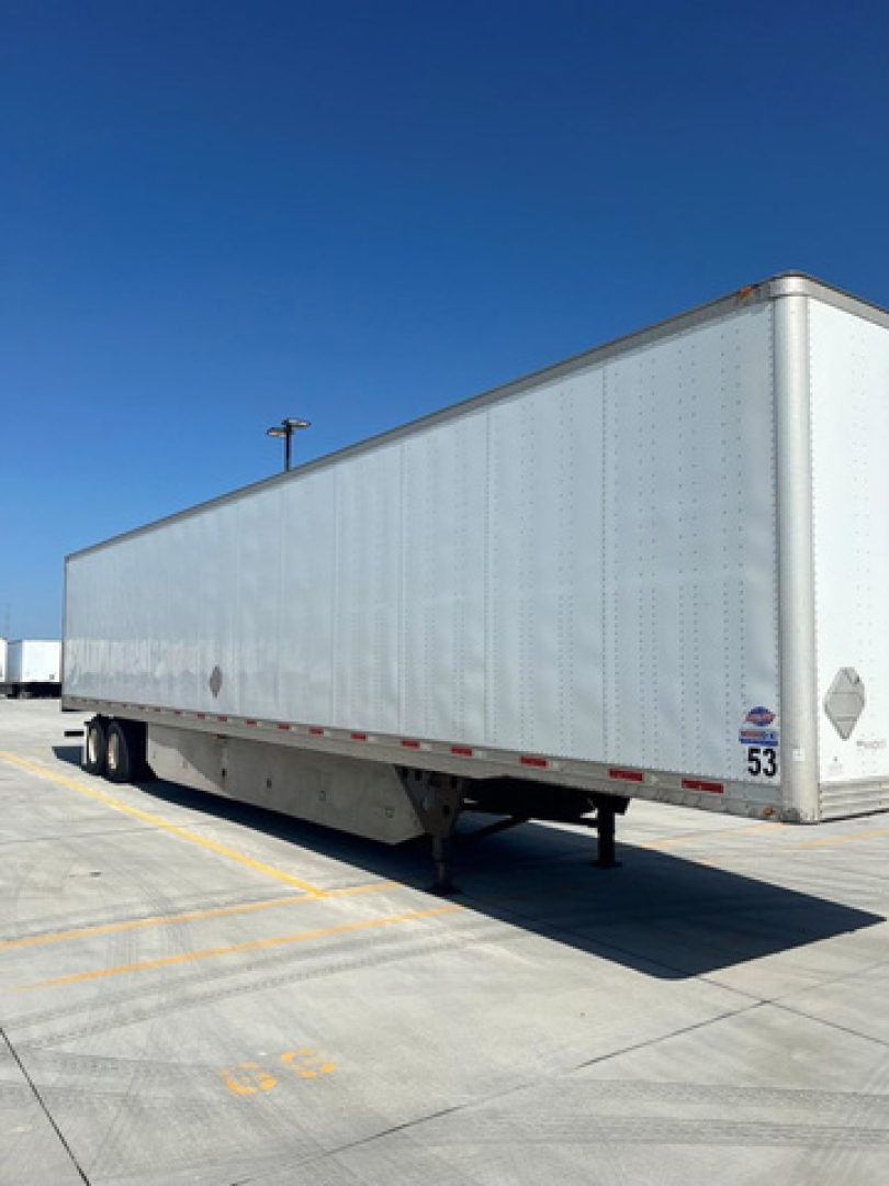Trailered utility van trailer with plain aluminum side, undercarriage shows two axles and support legs; no visible text on side apart from small reflective diamonds and numbers near rear.