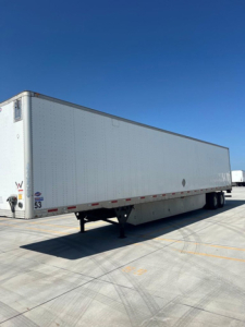 Utility van trailer on a concrete lot, side and front corners visible, under clear blue sky.