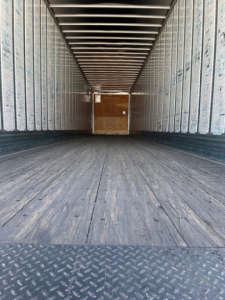 Trailer interior showing empty van with wooden bulkhead at far end, metal sidewalls, ribbed ceiling, and wooden planked floor.